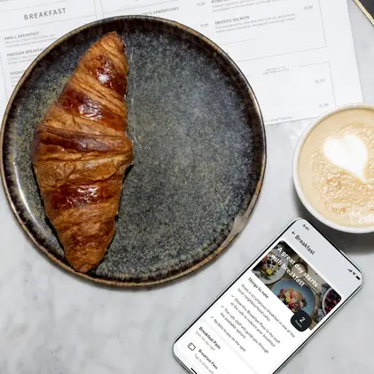 a croissant on a plate next to a cup of coffee and a phone