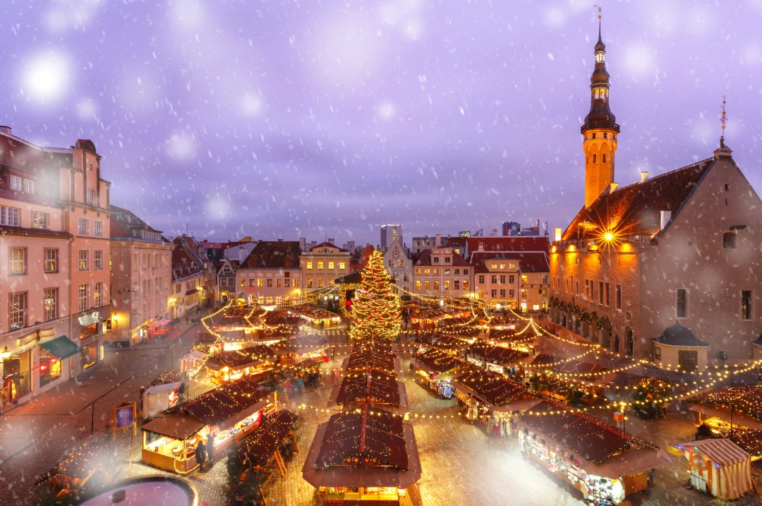 Snow falling on the Christmas market and tree on Tallinn's Town Hall Square, with the Town Hall tower lit up
