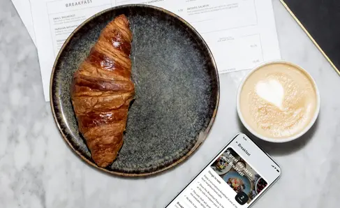 a croissant on a plate next to a phone and a cup of coffee