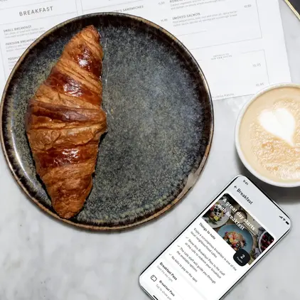 a croissant on a plate next to a phone and a cup of coffee