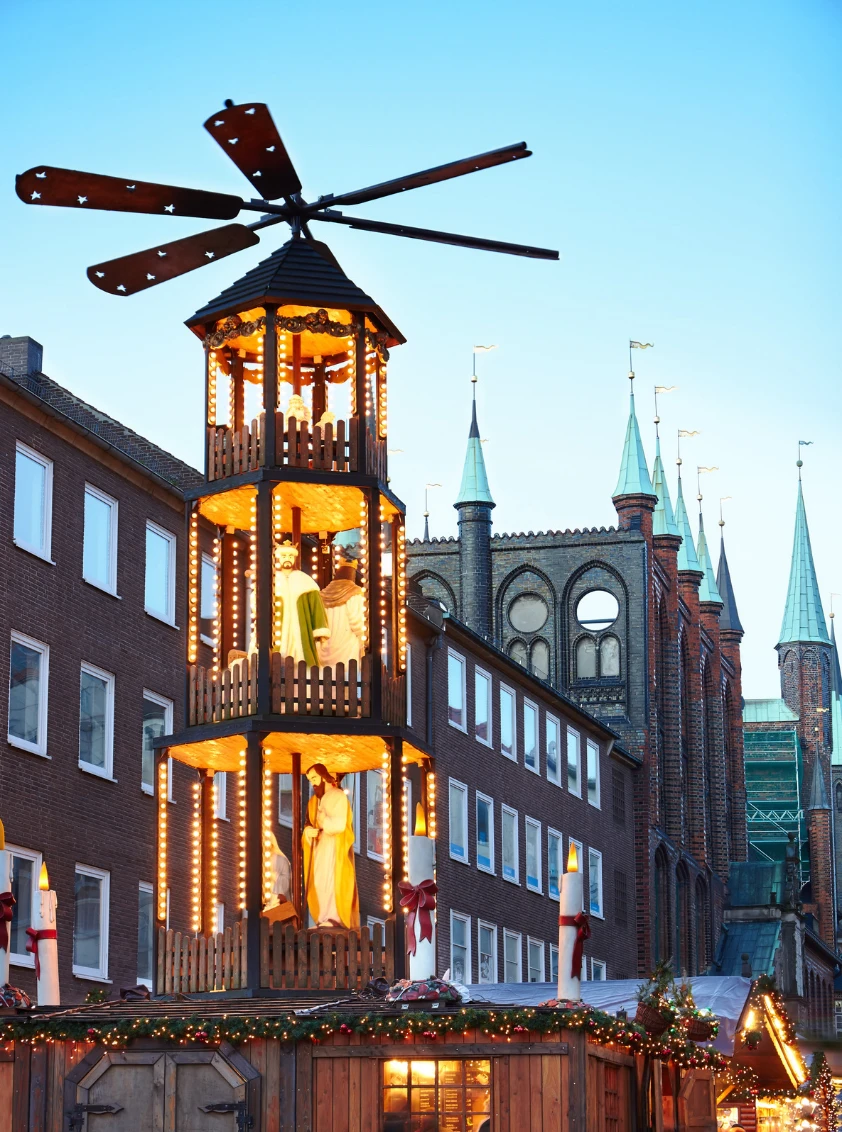 A wooden Christmas pyramid with nativity figures in front of the green spires of St Mary's Church, Lübeck