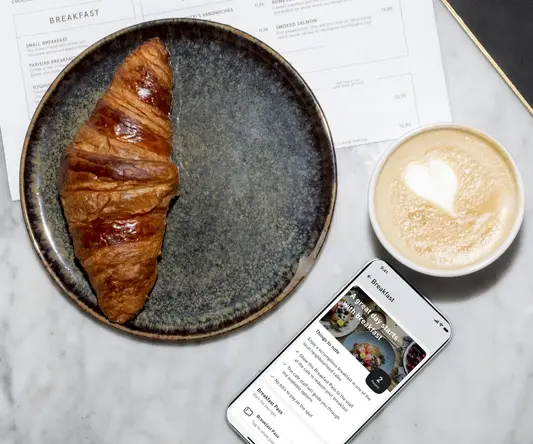 a croissant on a plate next to a cup of coffee and a phone